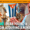 Cuban street scene with a large Cuban flag hanging between buildings; a man in a vest gestures toward the viewer beneath an orange banner.