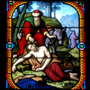 Stained-glass panel showing a bearded man in red and green kneeling beside a pale man lying on the ground, with a donkey behind them.