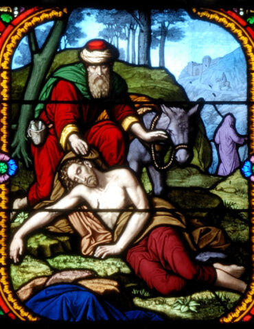 Stained-glass panel showing a bearded man in red and green kneeling beside a pale man lying on the ground, with a donkey behind them.