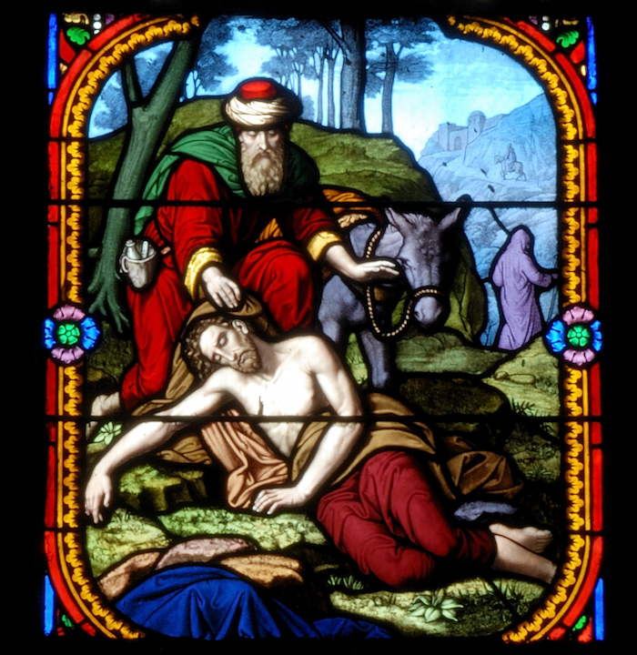 Stained-glass panel showing a bearded man in red and green kneeling beside a pale man lying on the ground, with a donkey behind them.